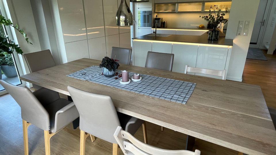 Essbereich mit hellem Holztisch, umgeben von beigefarbenen Stühlen und einer Kinderstuhlanordnung, angrenzend an eine moderne, offene Küche mit weißen Hochglanzschränken und einer beleuchteten Arbeitsplatte. Im Hintergrund sind eine große Fensterfront sowie Zimmerpflanzen sichtbar.