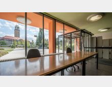 Blick durch große Fenster eines modernen Gebäudes mit einem langen Holztisch im Vordergrund auf eine Außenanlage mit gepflastertem Weg, Bäumen, Fahrrädern und einem historischen Turm mit rotem Dach und grüner Turmspitze im Hintergrund. Im Gebäude befindet sich zudem ein gläserner Eingangsbereich.