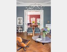 Wohnzimmer mit dunkelblau gestrichenen Wänden, modernen Möbeln und Holzfußboden; im Vordergrund ein niedriger Couchtisch mit Blumen und Büchern, dahinter gepolsterte Sessel und ein Design-Stuhl. Durch eine geöffnete Doppeltür ist ein angrenzender Raum mit roten Wänden, hängenden Lampen und einem Esstisch sichtbar.