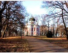 Pavillonähnliches Gebäude mit einer goldenen Kuppel und weiß-rosa Fassade, umgeben von kahlen Bäumen in einem parkähnlichen Gelände. Ein breiter Weg führt direkt zum Gebäude.