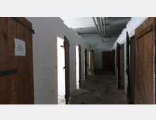 Langer Gang in einem Gebäude mit weißen Wänden, mehreren hölzernen Türen auf beiden Seiten und sichtbaren Rohren an der Decke. Auf einer der Türen hängt ein handgeschriebenes Schild mit der Aufschrift „KFZ“.