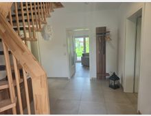 Heller Eingangsflur eines Wohnhauses mit großer Fliesenfläche, weiß gestrichenen Wänden und einer Holztreppe mit Geländer links. Rechts stehen eine schmale Garderobe und eine Laterne am Boden; durch eine offene Tür blickt man in einen Wohnbereich und weiter in den Garten.