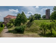 Historische Burganlage mit einem runden Turm aus Stein und einem Gebäude mit rotem Ziegeldach, umgeben von einer Grünfläche mit Bäumen und gepflegten Wegen. Im Hintergrund sind Menschen zu sehen, die sich auf einer erhöhten Terrasse aufhalten.