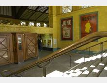Innenansicht eines Bahnhofsgebäudes mit gelblichen Wänden und großen Fenstern. Im Vordergrund ist ein Treppengeländer, im Hintergrund befinden sich Holztüren, historische Werbeplakate und Ticketautomaten.