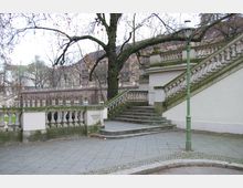 Steintreppe mit dekorativem Geländer in einem städtischen Park, umgeben von kahlen Bäumen und historischen Gebäuden im Hintergrund. Eine grüne Straßenlaterne steht auf der gepflasterten Fläche.