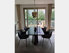 Essbereich mit einem rechteckigen Tisch aus schwarzem Glas und vier schwarzen Stühlen auf einem grauen Fliesenboden. Im Hintergrund große Fenster mit Blick auf eine Terrasse mit Holzplanken, Pflanzen und umliegender moderner Wohnbebauung.