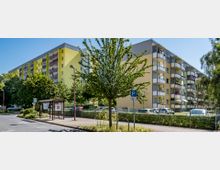 Mehrstöckiges Wohngebäude in hellgelber Farbe mit Balkonen, umgeben von Bäumen und parkenden Autos. Im Vordergrund befindet sich eine kleine Bushaltestelle mit Überdachung sowie ein Gehweg und ein Parkplatzschild.