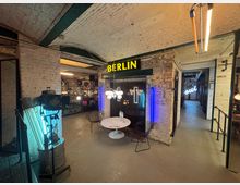 Industrie-inspirierter Innenraum mit weiß gestrichenen Backsteinwänden, sichtbaren Rohren und einer gelben Leuchtschrift mit der Aufschrift „BERLIN“. Im Vordergrund stehen ein runder Tisch und Stühle, umgeben von modernem Dekor und neonbeleuchteten Elementen.