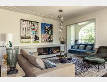 Wohnzimmer mit heller, moderner Einrichtung, beige Sofas, blauen Kissen und dunklem Sessel. An der Wand befinden sich gerahmte Kunstwerke, im Hintergrund ist eine Nische mit einem blauen Sofa vor einem großen Rundbogenfenster zu sehen, das Blick ins Grüne bietet.