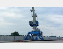 Blauer Hafenkran auf einer betonierten Fläche vor einer hohen Betonmauer; im Hintergrund sind Bäume und kleine Gebäude mit grünen Dächern sichtbar. Der Kran ist mit einer Kabine und einem hohen Ausleger ausgestattet.