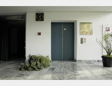 Ein Gebäudeflur mit einer grauen Marmorbodenfläche, in dessen Mitte ein geschlossener blauer Aufzug zu sehen ist. An der weißen Wand hängen eine analoge Wanduhr, ein Flucht- und Rettungsplan sowie ein Feueralarm, während grüne Topfpflanzen den Bereich dekorieren.