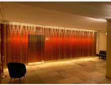 Flur mit glänzenden, durchscheinenden Vorhängen in Gelb-Gold-Tönen, die vor einer roten Wand hängen. Der Raum hat eine helle Deckenbeleuchtung, Fliesenboden und Sitzmöbel, darunter ein schwarzer Sessel und eine schlichte schwarze Bank.