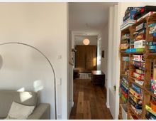 Wohnzimmer mit Blick auf einen Flur. Links ein graues Sofa mit einer geschwungenen Stehlampe, rechts ein Regal mit Brettspielen, im Hintergrund Holzdielenboden und ein Schrank aus Holz.