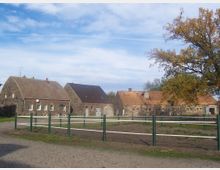 Ein ländlicher Hof mit mehreren Gebäuden aus Naturstein, Giebeldächern und roten Ziegeln. Im Vordergrund befindet sich ein leerer, umzäunter Bereich mit einem Baum und Kopfsteinpflasterweg.