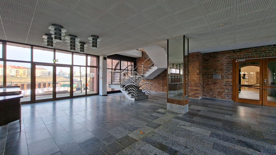 Großzügige, moderne Eingangshalle mit grauem Steinboden, Backsteinwänden und einer zentralen Treppe mit Edelstahlgeländer. Links sorgen bodentiefe Glasfronten für Tageslicht, rechts führen Glastüren in angrenzende Räume; an der Decke hängen runde Leuchten.