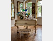 Ein klassischer hellbeiger Flügel mit goldenen Verzierungen steht in einem elegant eingerichteten Raum mit Parkettboden. Auf dem Flügel befinden sich Geschenke, Luftballons und Blumenarrangements; im Hintergrund sind hohe Fenster mit gelben Vorhängen sowie vergoldete Spiegel und Gemälde an den Wänden zu sehen.