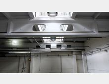 Industriehalle mit weiß gestrichenen Wänden und Deckenbalken. Sichtbare Rohrleitungen, hängende Leuchtstofflampen und ein Oberlicht mit Metallverstrebungen prägen die industrielle Architektur.