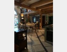 Gemütlicher Wohnraum mit Holzbalkendecke, gemustertem Fliesenboden und einem langen Esstisch mit verschiedenen Stühlen. Im Hintergrund sind große Fenster mit Blick auf den Außenbereich, eine hölzerne Galerie und rustikale Möbel zu sehen.