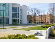 Moderne Gebäude mit weißen Fassaden und großen Glasflächen links, daneben ein historisches Backsteingebäude mit Ziergiebeln rechts. Im Vordergrund gepflasterte Wege, Grünflächen und ein niedriges Geländer.