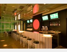 Eine modern gestaltete Bar mit einer langen, gemusterten Theke, schwarzen Barhockern und beleuchteten Elementen. Im Hintergrund sind Regale mit Flaschen, eine Kaffeemaschine sowie Grün- und Rotakzente auf Wänden und Decken sichtbar.