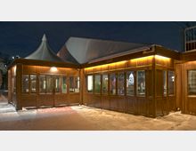 Holzgebäude mit großen Fenstern und angeschriebenem "Bar"-Bereich, beleuchtet mit warmem Licht, bei Nacht. Im Hintergrund ist eine Zeltstruktur sichtbar, der Boden ist mit Schnee bedeckt.