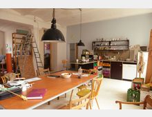 Ein heller Raum mit einer offenen Küche und einem Essbereich. Ein großer Holztisch mit verschiedenen Stühlen steht im Vordergrund, während Regale, Küchengeräte und offene Ablagen die Küche im Hintergrund prägen.