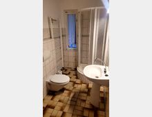 Kleines Badezimmer mit beigefarbenen Fliesen an den Wänden und braun-gemusterten Fliesen auf dem Boden. Das Zimmer verfügt über eine Toilette, ein Waschbecken, eine Duschecke mit Glastür sowie einen Handtuchheizkörper und ein schmales Fenster mit weißer Spitzen-Gardine.