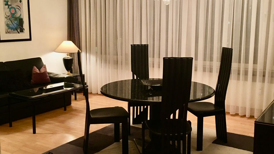 Modernes Wohn- und Esszimmer mit rundem schwarzem Esstisch und vier hohen Stühlen auf einem karierten Teppich, darüber ein großer Kristalllüster. Im Hintergrund bodentiefe Fenster mit weißen Vorhängen, links ein schwarzes Sofa mit Couchtisch, Stehlampe und gerahmtem Wandbild.