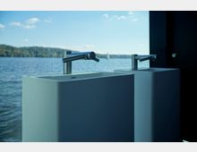 Zwei moderne freistehende Waschbecken mit silbernen Wasserhähnen vor einer großen Glasfront, die den Blick auf einen See mit Wald im Hintergrund und einem segelnden Boot bietet.
