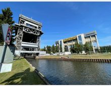 Ein moderner Wasseraufzug aus Stahl mit Gitterstruktur steht neben einem Wasserkanal, dahinter ein großes, gelb-grau akzentuiertes Verwaltungsgebäude mit Glasfassade. Der Hintergrund zeigt grüne Bäume und einen blauen Himmel.