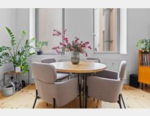 Heller Wohnraum mit einem runden Holztisch, umgeben von gepolsterten Stühlen. Im Hintergrund sind große Fenster, Zimmerpflanzen und ein orangefarbenes Regal mit Büchern zu sehen, der Boden besteht aus hellem Holz.