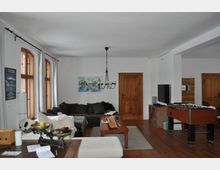 Wohnzimmer mit Holzboden, weißen Wänden und mehreren Holztüren. Der Raum enthält eine große dunkle Eckcouch, einen Holztisch, einen Tischkicker und eine Wanddekoration mit Gemälde, sowie einen Kronleuchter an der Decke. Drei große Fenster mit Holzrahmen lassen Licht herein.