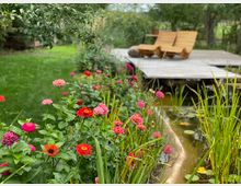 Ein Garten mit bunten Blumen wie roten, rosa und orangen Zinnien rechts neben einem natürlichen Teich mit Schilf und Seerosen. Im Hintergrund steht ein Holzsteg mit zwei hölzernen Liegestühlen, umgeben von grünem Rasen und Bäumen.