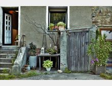 Eine Hausfassade mit einer offenen weißen Tür auf der linken Seite, mehreren blühenden Pflanzen in Töpfen vor einem Fenster, sowie einem alten Holztisch und einem verwitterten Holztor auf der rechten Seite. Die Umgebung wirkt rustikal, mit sichtbaren Putz- und Backsteinflächen.