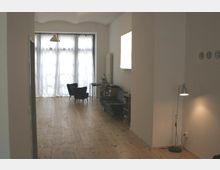 Heller Wohnraum mit Holzboden, schlichten weißen Wänden und einer Fensterfront mit transparenten Vorhängen im Hintergrund. Der Raum ist spärlich möbliert mit einem Sessel, einer Stehlampe und kleinen Tischen.