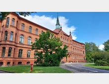 Backsteingebäude im historischen Stil mit großen Rundbogenfenstern und einem spitzen grünen Turm, umgeben von Grünflächen und einem gepflasterten Weg im Vordergrund. Der Himmel ist teils bewölkt mit blauem Himmel.