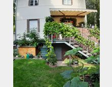 Wohnhaus mit Garten: Eine Treppe führt zu einer überdachten Veranda mit Pflanzen und rot blühenden Geranien an einem weißen Gebäude mit Backsteinelementen. Im Vordergrund befinden sich eine Rasenfläche, Gemüsepflanzen, Gießkannen und ein Holzschuppen.