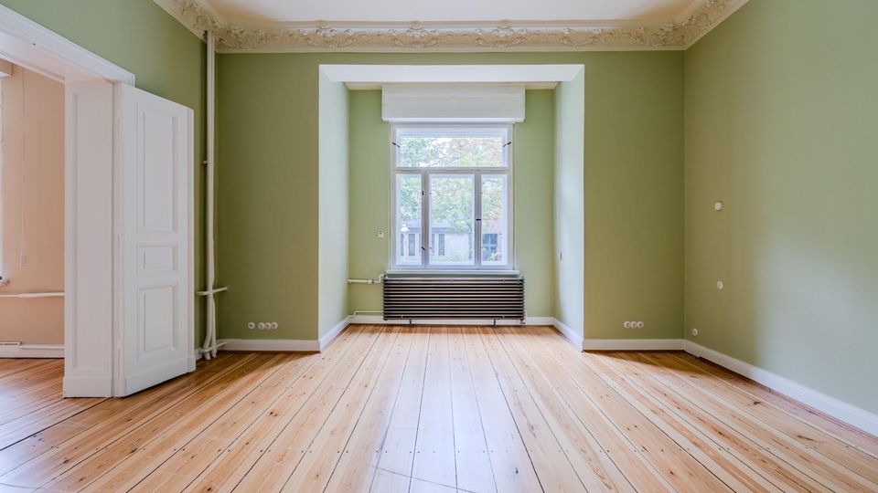 Leerer Wohnraum mit hellgrünen Wänden, Holzdielenboden und weißer Stuckdecke. In der Mitte ein großes Fenster in tiefer Nische mit Heizkörper darunter sowie eine offene weiße Flügeltür zum Nebenraum.