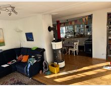 Helles Wohnzimmer mit Holzboden und einem schwarzen Ecksofa, auf dem Decken und Kissen liegen. Ein weißer Kamin trennt den Wohnbereich von der angrenzenden Küche mit Esstisch und Stühlen; bunte Girlanden hängen über dem Durchgang.