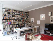 Büro- oder Arbeitszimmer mit einer großen Bücherwand voller Bücher, einem weißen Schreibtisch mit Computer und Schreibtischstuhl. An der braunen Wand hängen mehrere gerahmte Bilder, daneben steht ein Fernseher auf einem kleinen Regal, und im Vordergrund ist ein gemusterter Sessel mit einer bunten Decke zu sehen.