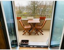 Kleiner Balkon mit Holzboden, zwei klappbaren Holzstühlen und einem Tisch. Im Hintergrund sind ein Geländer, Blumenkästen sowie kahle Bäume und eine urbane Umgebung sichtbar.