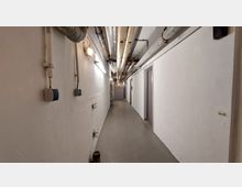 Schmaler, funktionaler Korridor mit weißen Wänden und grauem Boden, an der Decke verlaufen zahlreiche freiliegende Rohre und Leitungen. Mehrere Türen befinden sich entlang der rechten Wand, und der Korridor endet an einer geschlossenen Tür.