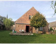 Backsteinbauernhaus mit steilem Giebeldach, einer kleinen Holzterrasse und einem Baum im Vorgarten. Im Hintergrund sind weitere ländliche Gebäude sowie die Spitze eines Kirchturms sichtbar.