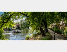 Flusslandschaft mit einem kleinen Ausflugsschiff auf dem Wasser, umgeben von Bäumen und Grünflächen; rechts führt ein gepflasterter Weg entlang einer Backsteinmauer, gesäumt von Laternen.