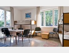 Schlicht eingerichtetes Zimmer mit Etagenbetten, vier Stühlen um einen rechteckigen Tisch, einem kleinen Holztisch und großen Fenstern, die Tageslicht hereinlassen und den Blick auf Bäume im Außenbereich ermöglichen. Auf dem Boden liegen bunte Teppiche, und an der Wand hängt ein gerahmtes Bild.