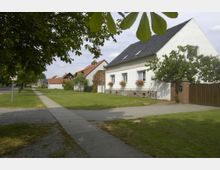 Straßenszene in einem Dorf mit weißen Wohnhäusern, Vorgärten und Gehweg; das vordere Haus hat ein dunkles Schrägdach, Blumenkästen an den Fenstern und wird von einem Baum sowie einer Hecke umgeben. Im Hintergrund sind weitere Gebäude mit roten Ziegeldächern zu sehen.