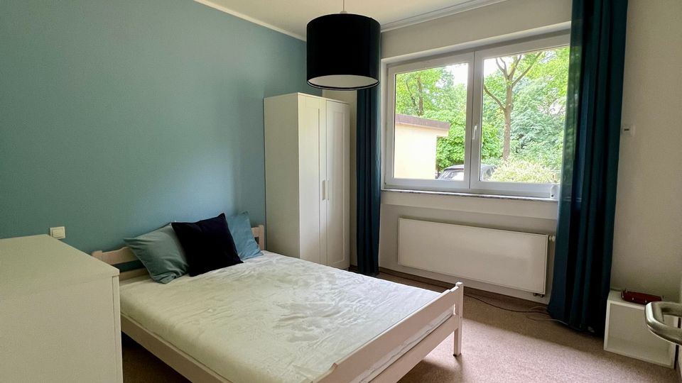 Helles Schlafzimmer mit weißem Bettgestell, Kleiderschrank und Kommode, einer blau gestrichenen Akzentwand und Teppichboden. Großes Fenster mit dunklen Vorhängen und Heizkörper darunter, Blick ins Grüne.