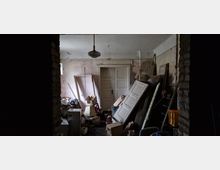 Unaufgeräumter, dunkler Raum mit einer alten Holztür im Hintergrund. Der Raum ist gefüllt mit Möbeln, Kartons, einer losen Tür und verschiedenen Baumaterialien, die stapelweise gelagert sind; eine Hängelampe hängt von der Decke.