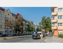 Straßenszene mit mehrstöckigen Wohnhäusern im Altbaustil, beidseitig gesäumt von Bäumen. Autos parken entlang der Straße, und im Vordergrund biegt der Gehweg um eine Ecke, während ein Straßenschild und eine Werbetafel am Straßenrand stehen.