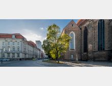 Straßenszene mit einer historischen Backsteinkirche auf der rechten Seite, gekennzeichnet durch hohe Spitzbogenfenster, und einem großen Baum in der Mitte des Platzes. Links steht ein klassizistisches Gebäude mit rotem Dach, und im Hintergrund ist ein modernes Hochhaus sichtbar.